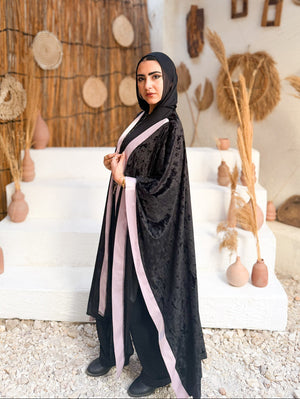 Woman in a black abaya with pink accents standing in a rustic outdoor setting.