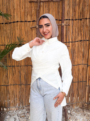Woman wearing a hijab and white shirt standing in front of a woven wall with a plant to her left.