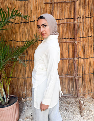 Woman in a white outfit standing in front of a woven reed wall with a plant to her left.