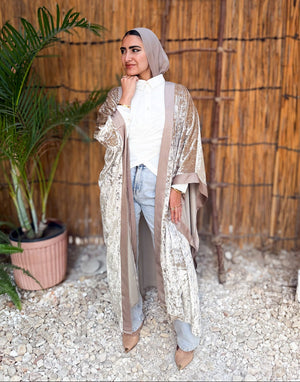 Woman in a long beige coat standing in front of a woven wall with a plant to her left.