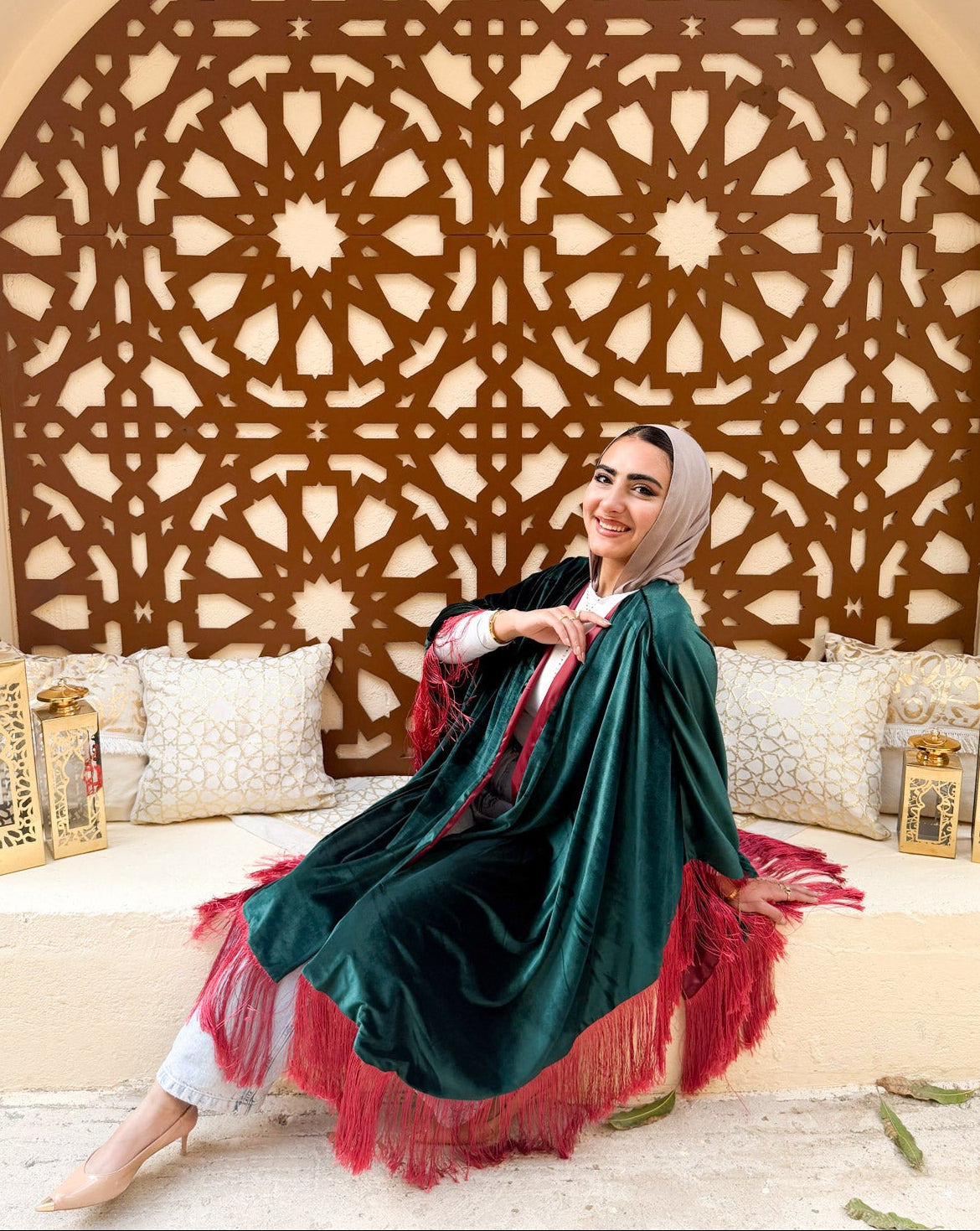 Woman in a green and red traditional outfit sitting in a decorated room with intricate wall patterns.