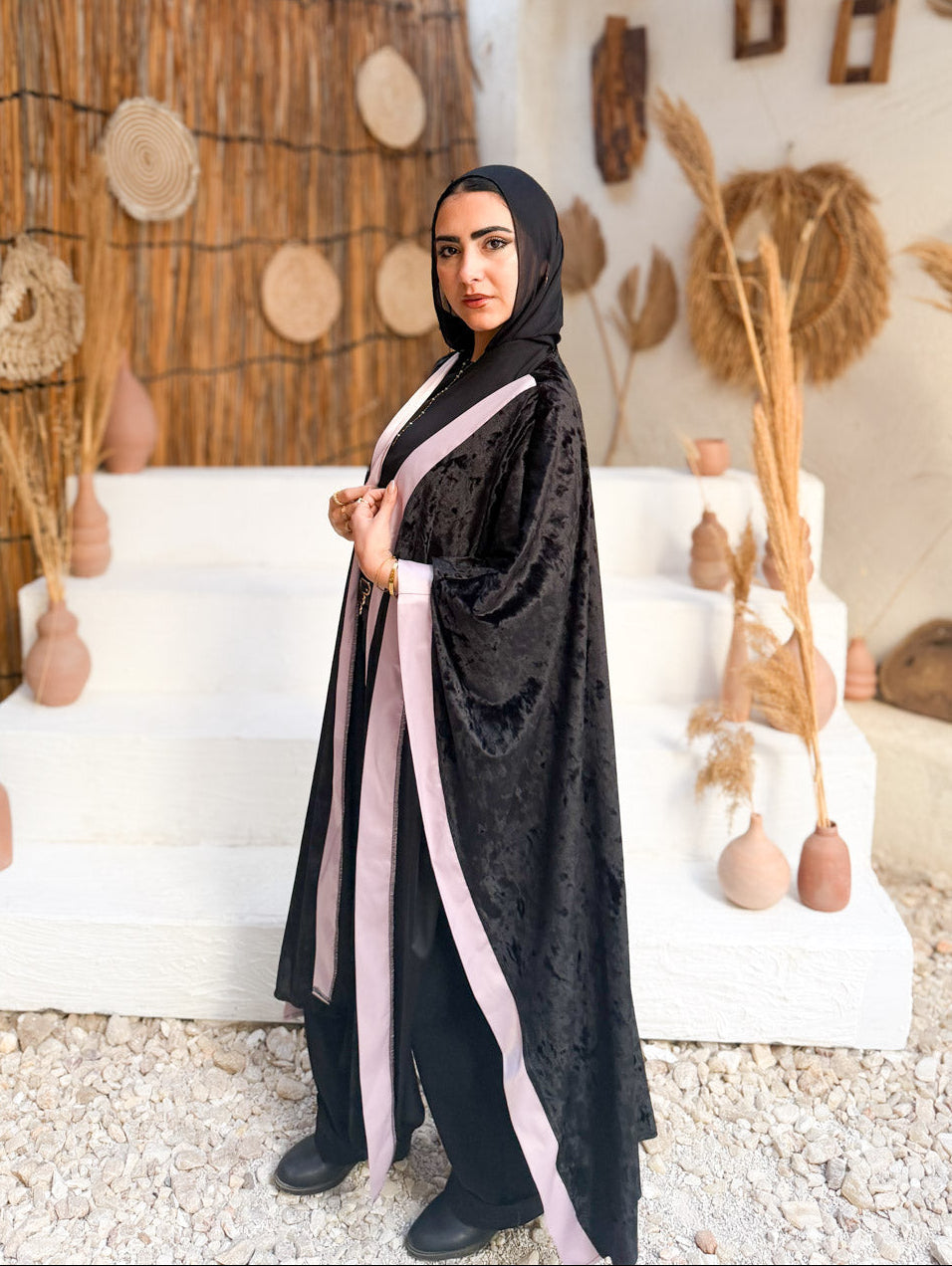 Woman in a black abaya with pink accents standing in a rustic outdoor setting.