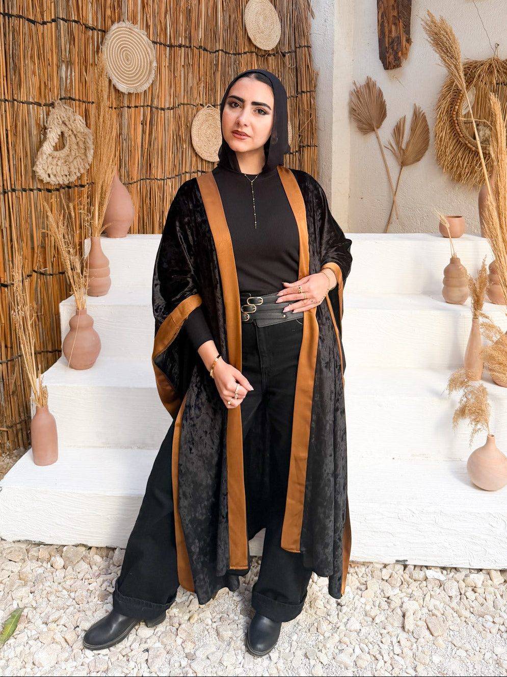 Woman in a black and brown outfit standing in a decorative outdoor setting with straw walls and decor.