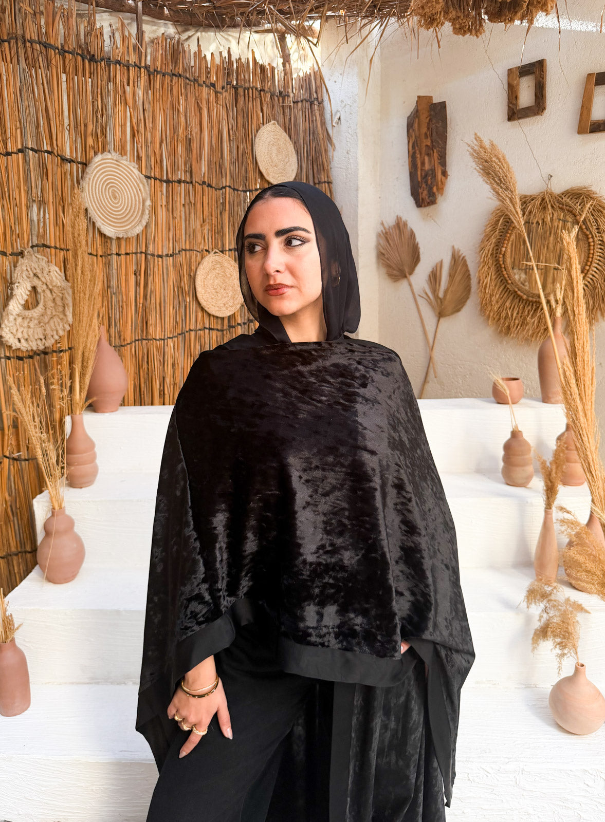 Woman wearing a black hooded garment in a decorative setting with woven wall and dried plants.