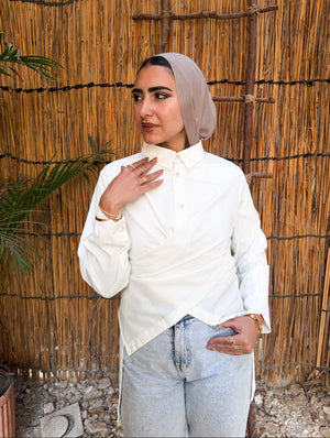 Woman wearing a beige hijab and white blouse against a reed wall.