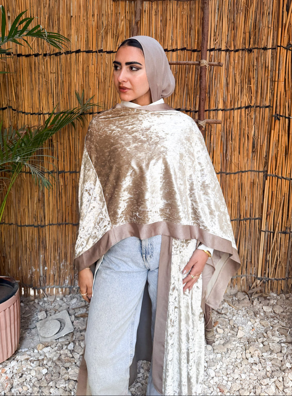 Woman wearing a gold shawl and light blue pants standing against a thatched wall with plants around.